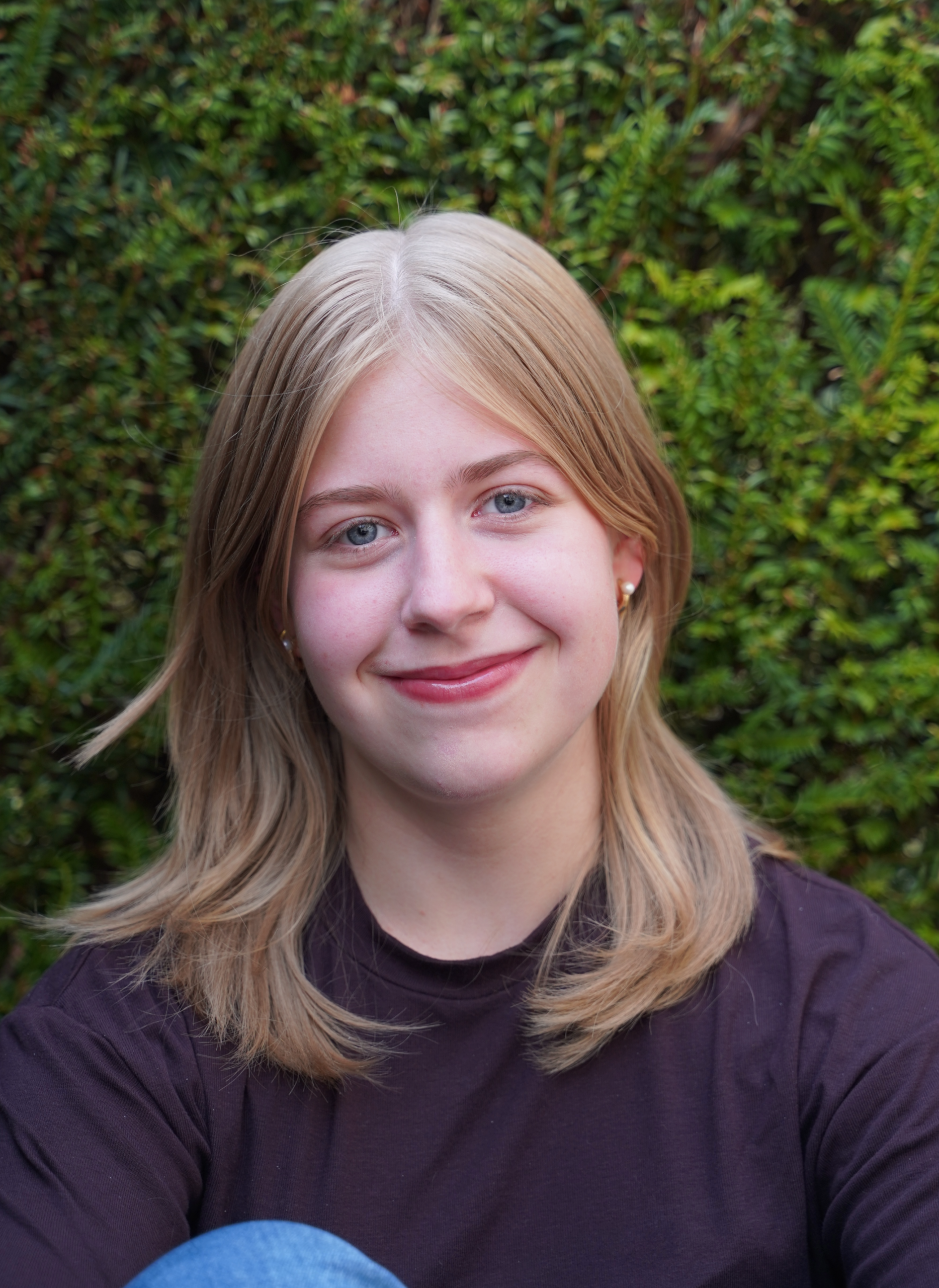 Eine junge Frau mit schulterlangem, blonden Haar und braunem Oberteil posiert vor einer Hecke. Sie lächelt in die Kamera.